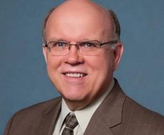 Smiling man in glasses and brown suit against blue background.