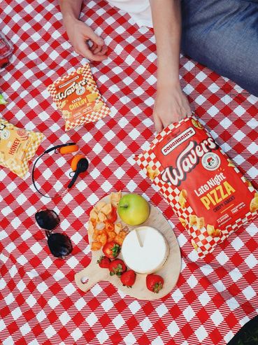 Hand reaching for pizza-flavored Wafers on a red checkered picnic blanket with fruit and cheese.