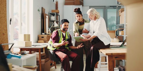 Three coworkers discussing plans in a bright office with boxes and shelves.