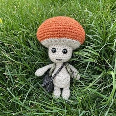 Crocheted mushroom character with a bag sitting on grass.