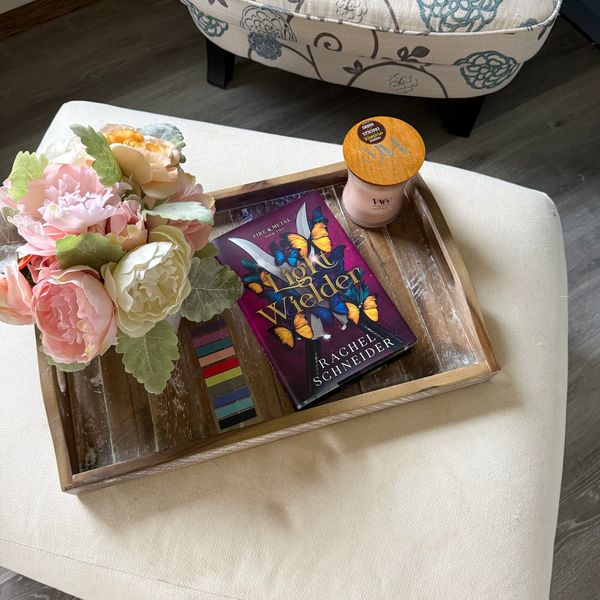 Wooden tray with book, candle, and flowers on white surface.