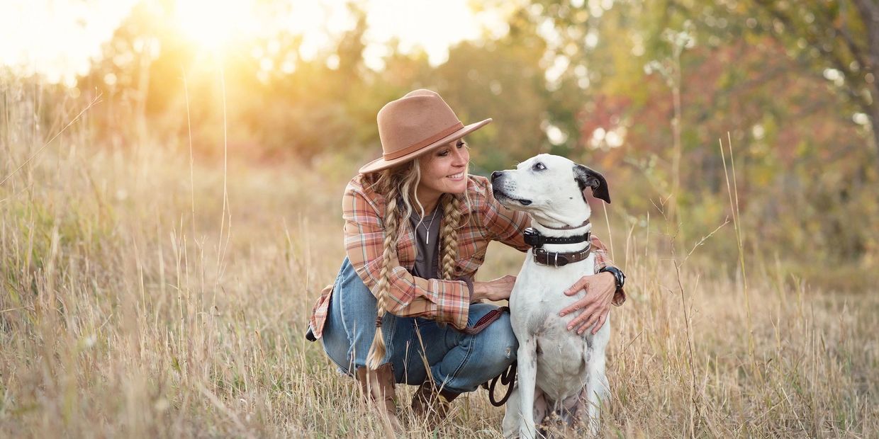 Woman in a hat lovingly hugging her white dog in a sunlit field.