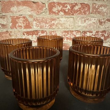 Five amber ribbed glass candle holders on a dark surface against a brick wall.