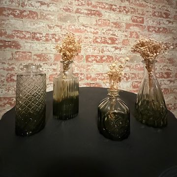 Four decorative glass vases with dried flowers on a black table against a brick wall.