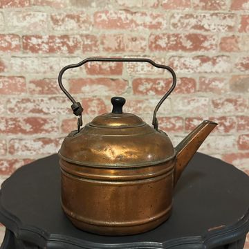 Vintage copper kettle on a black ornate table against a brick wall.