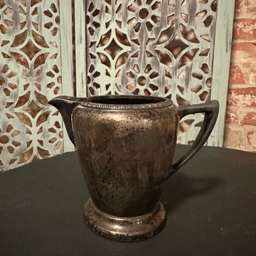 A tarnished silver pitcher on a dark surface with a decorative backdrop.