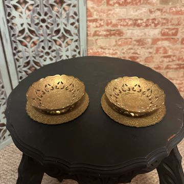 Two ornate golden bowls on a black round table with a decorative screen and brick wall background.