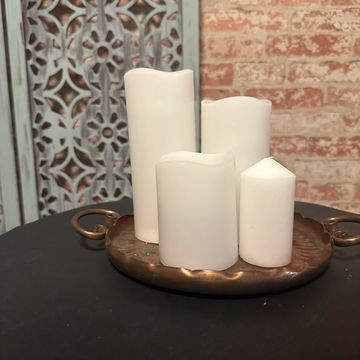 Four white candles of varying shapes on a bronze tray against a brick wall backdrop.