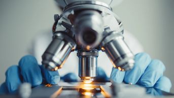 Photograph of a 4-lens microscope and gloved hands--a lab worker is directly behind the microscope.