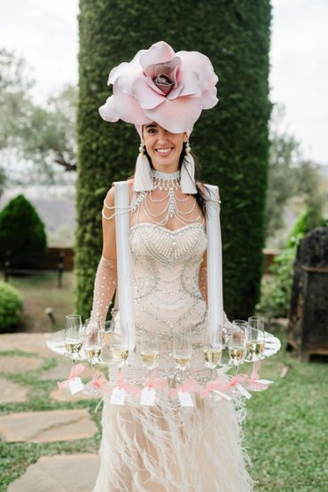 Woman wearing an extravagant floral hat and a pearl-embellished dress serving champagne.