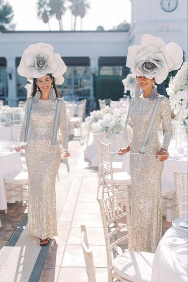 Two women in sparkling dresses and oversized white flower hats at a bright outdoor event.