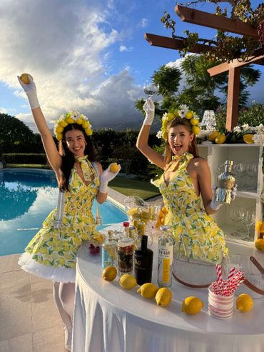 Two women in lemon-themed dresses celebrate by a pool with lemons and drinks.