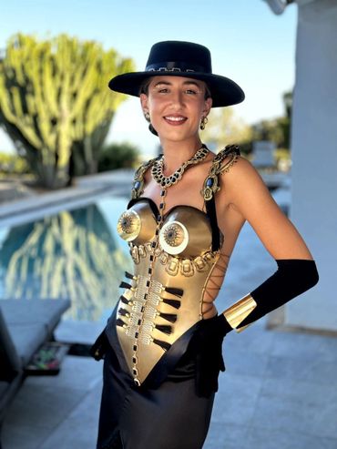 Woman in a striking gold corset, black hat, and gloves posing by a pool.