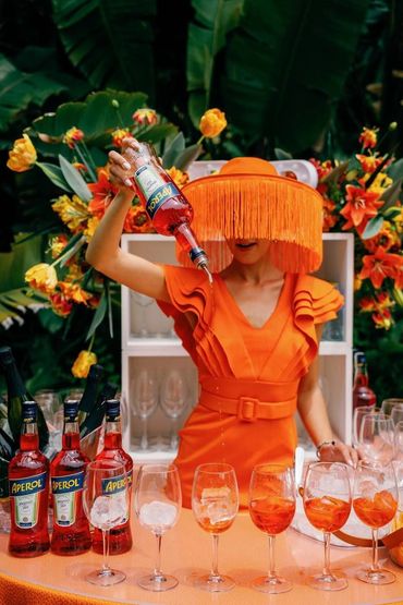 Woman in vibrant orange dress and hat pouring Aperol into glasses at a lively floral bar setup.
