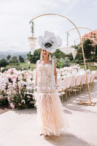 Woman in elegant feathered dress and giant flower hat holding glasses at outdoor event.