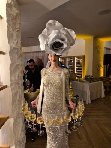 Woman in sparkling gown with a giant flower hat serving champagne glasses.
