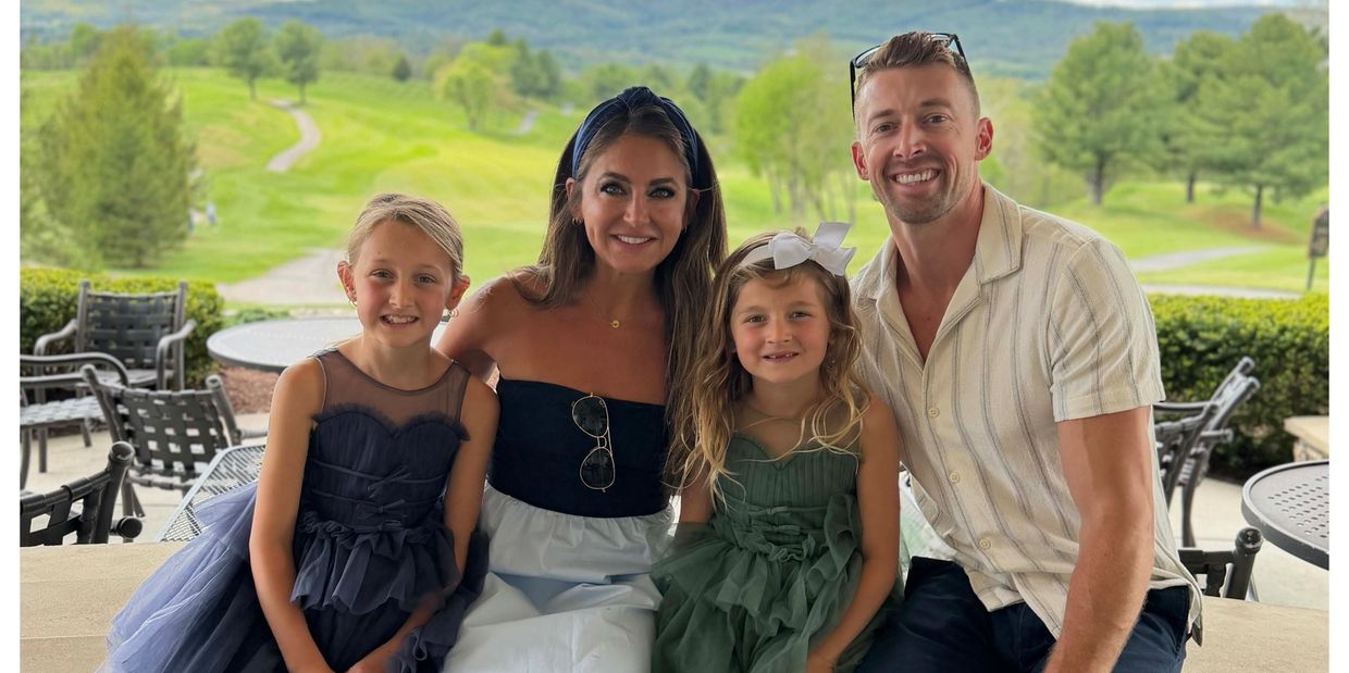 A family of four smiling outdoors with a scenic green background.