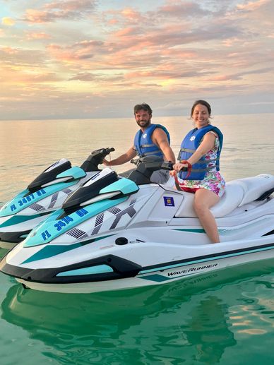 A smiling couple on jet skis at sunset over calm waters.