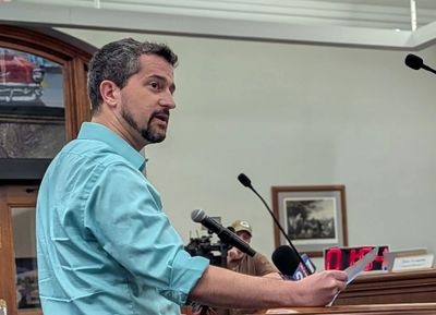 Man in teal shirt speaking at a podium with microphones in a formal setting.
