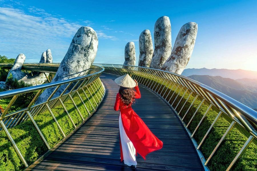 Woman in red dress and conical hat on a bridge held by giant stone hands.