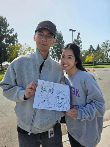 Asian couple in their early twenties smiling and together holding up their caricature drawing.