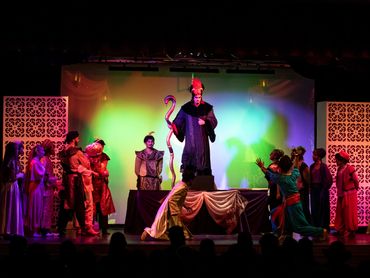 Actors perform a vibrant theatrical scene with a central figure in a tall hat on stage.