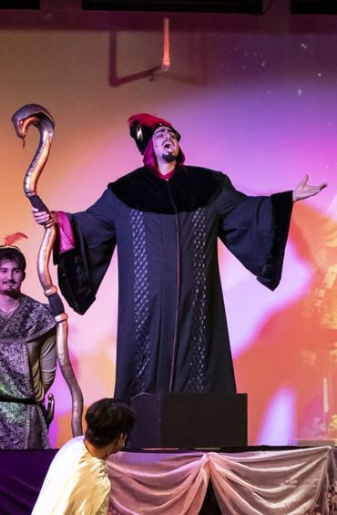 Actor in black robe and red headdress performing on stage with a large staff.