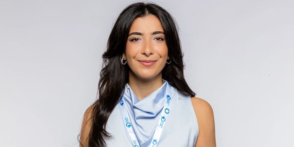 Young woman in a light blue top and jeans with a conference badge smiling.