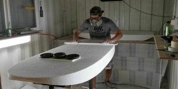 Man shaping a large foam surfboard in a workshop.