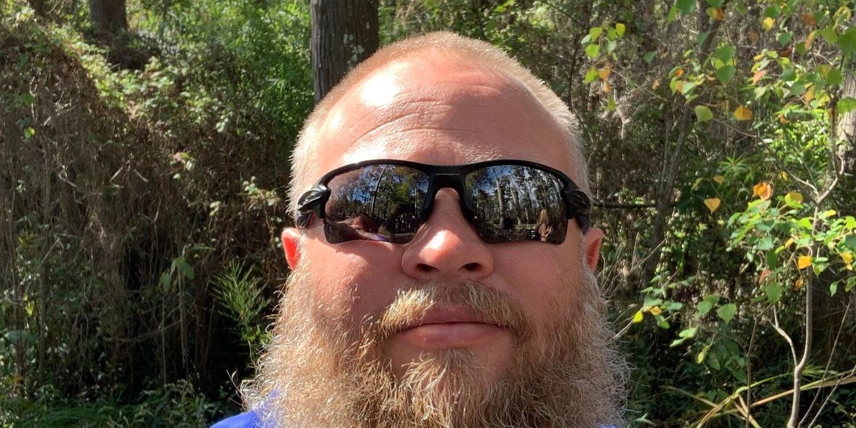 Man with a beard and sunglasses in a forest setting on a sunny day.