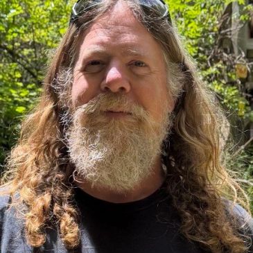 Man with long curly hair and a full beard outdoors in sunlight.