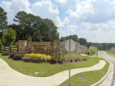 Entrance sign for Alcovy Creek neighborhood with houses and sidewalks under a partly cloudy sky.