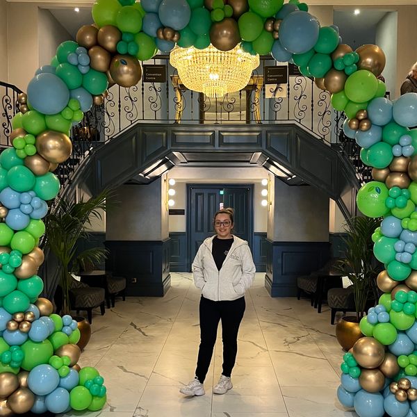 Person posing under a colorful balloon arch in an indoor setting.