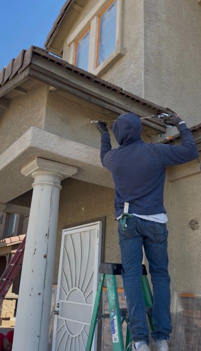 Person in blue hoodie fixing roof gutters on a house using a ladder.