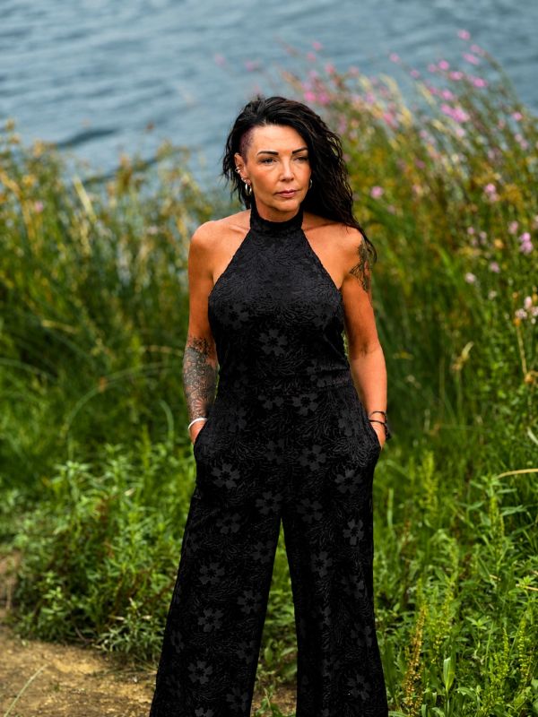 Woman in black floral jumpsuit near water and greenery.