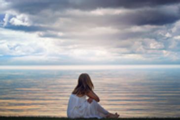 Woman in white dress sitting on a beach looking at the water.