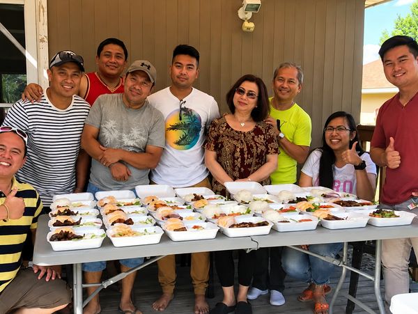 Group of nine people standing behind a table full of food containers outdoors.