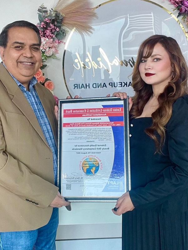 Two people holding a framed certificate in a decorated makeup and hair studio.
