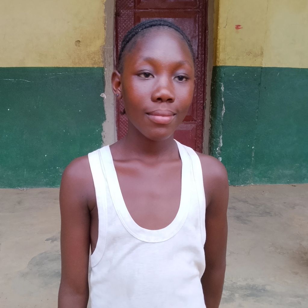 A young girl in a white tank top stands in front of a green and yellow wall.