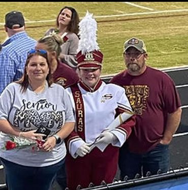 A young marching band member in uniform stands with family at a sports event.