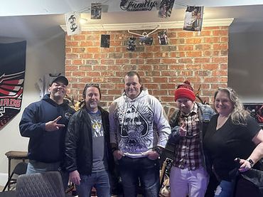 Five friends posing indoors in front of a brick wall with family photos.