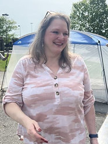 Smiling woman in a light pink patterned shirt outdoors near a tent.