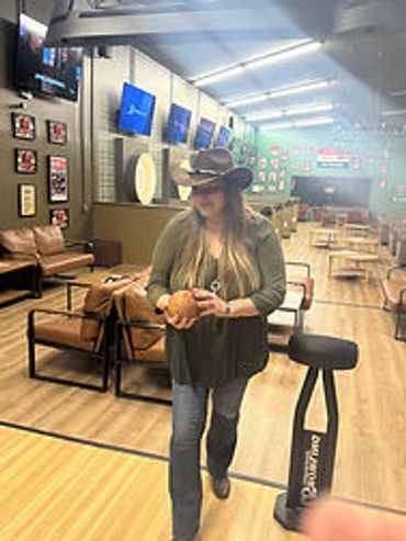 Woman in cowboy hat walking in a bowling alley carrying a bowling ball.