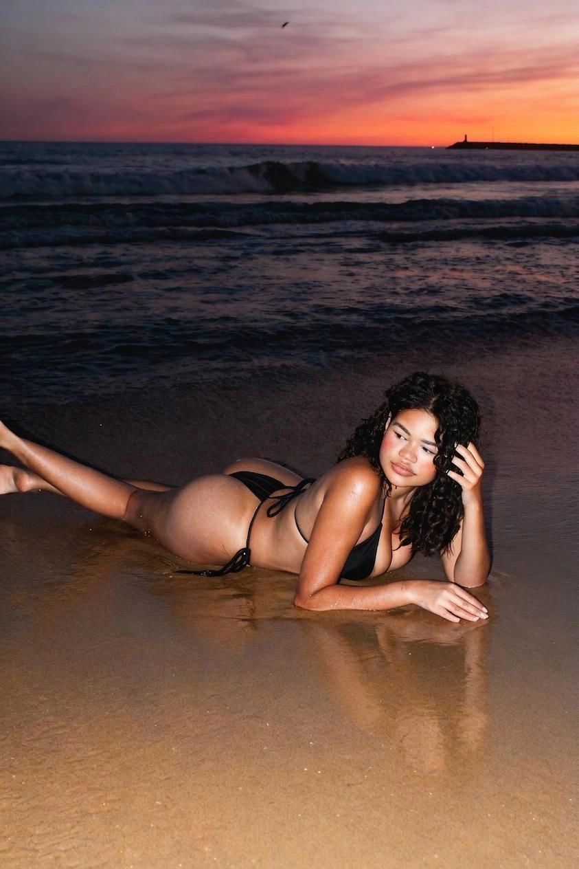 Woman in black bikini lying on wet sand at sunset on the beach.