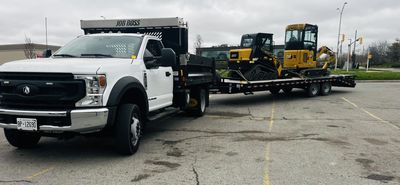 White flatbed truck towing two yellow construction vehicles on a trailer.