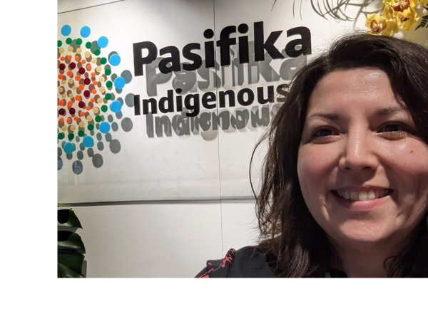 A woman smiling next to a wall sign that reads 'Pasifika Indigenous'.