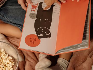 Child and adult reading a picture book with popcorn nearby.