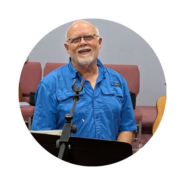 Smiling elderly man in blue shirt standing behind a microphone stand.