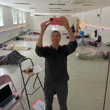 A woman takes a selfie in a yoga class with participants lying on mats in the background.