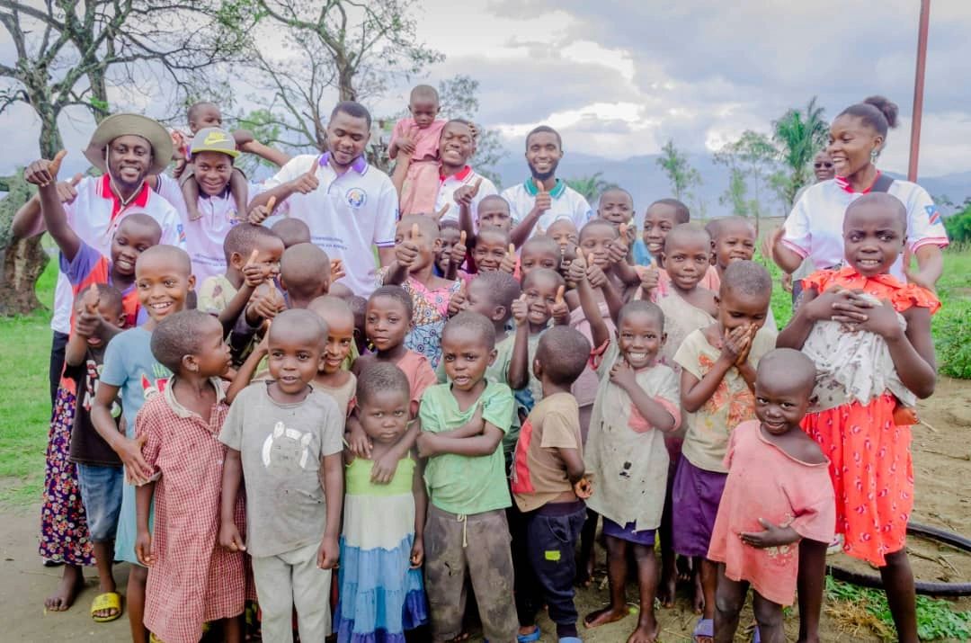 Group of smiling children and adults outdoors giving thumbs up.